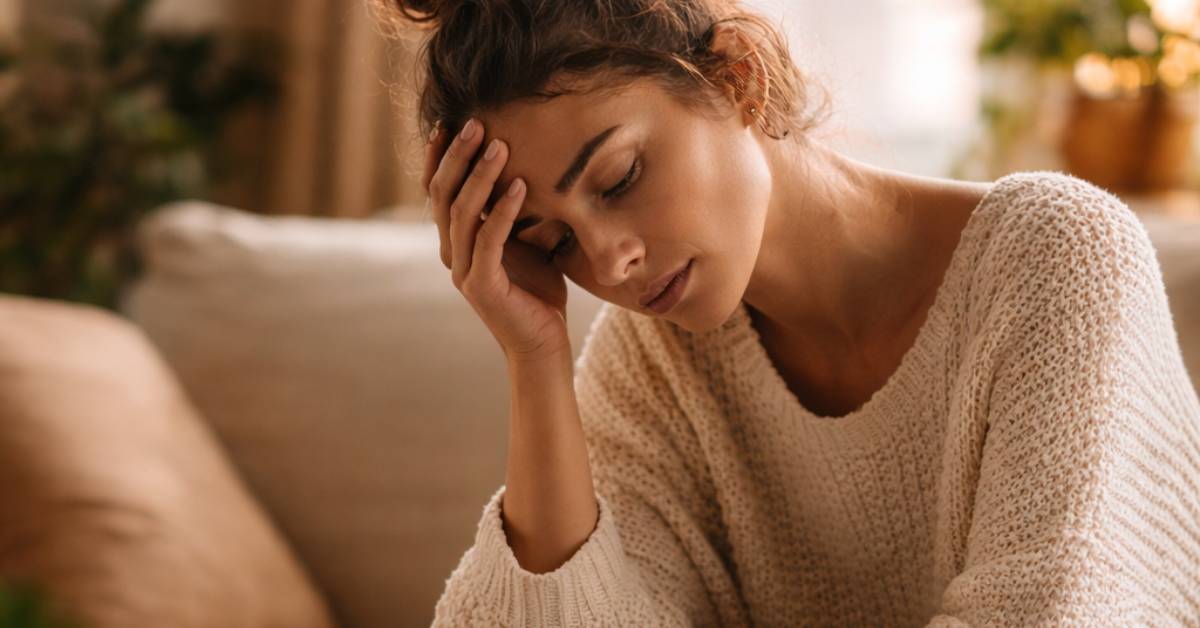 Femeie asezata pe canapea, cu mana la frunte, intr-un spatiu cald si linistit